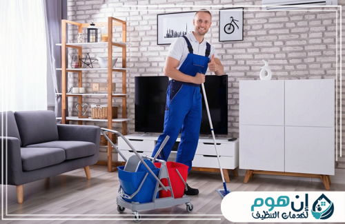 شركة تنظيف كنب بجدة 0559016158 شركة إين هوم للتنظيف عامل نظافة مبتسم يحمل ممسحة داخل صالة مجهزة بأدوات التنظيف.